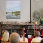 Dwie kobiety przemawiają do publiczności w bibliotece. Za nimi wyświetlany jest duży plakat dotyczący wydarzenia literackiego. W tle widoczne są półki z książkami i rośliny doniczkowe.