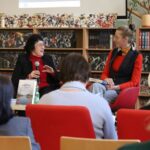 Dwie kobiety siedzą i rozmawiają przed półkami z książkami podczas wydarzenia dyskusyjnego; jedna trzyma mikrofon, podczas gdy druga słucha, a kilka osób siedzi na widowni, a książki są wyświetlane na pobliskim stole.