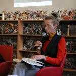 Kobieta w okularach i czerwonym swetrze siedzi na czerwonym krześle, trzymając mikrofon i czytając z gazet. Za nią znajdują się półki wypełnione komiksami, a powyżej widoczny jest wyświetlany slajd z wydarzenia.