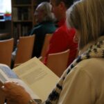 Osoba z siwymi włosami i pasiastym szalikiem czyta książkę, siedząc między innymi w pokoju z drewnianymi krzesłami i regałami.