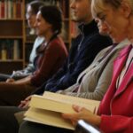 Cztery dorosłe osoby siedzące w rzędzie w bibliotece, za nimi półki z książkami. Osoba najbliżej kamery, ubrana w różową kurtkę, czyta książkę. Pozostali uważnie patrzą przed siebie.