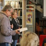 Mężczyzna trzymający mikrofon i książkę przemawia do publiczności w bibliotece, podczas gdy kobieta stoi w pobliżu. W tle widoczne są półki z książkami i plakaty.