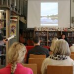 Grupa ludzi siedzi przed ekranem w bibliotece, oglądając prezentację. Mężczyzna stoi czytając książkę, podczas gdy dwie kobiety siedzą przy stole przed regałami. W tle widoczne są rzędy książek i schody.