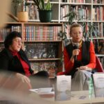 Dwie kobiety siedzą w bibliotece, jedna z nich trzyma mikrofon podczas przemowy. Książki i rośliny doniczkowe wypełniają półki za nimi. Kilka książek i butelka wody leżą na stole przed nimi.