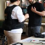 Kobieta w czarnej kamizelce rozmawia z mężczyzną w czarnej koszuli przy stole z różnymi przedmiotami i dokumentami w pokoju z oprawionymi obrazami na ścianach.