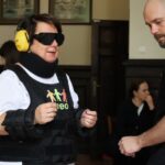 Kobieta nosząca czarne gogle, żółte ochraniacze na uszy, usztywnienie szyi i wyściełany sprzęt jest wspomagana przez mężczyznę. Dwie osoby siedzą w tle w pokoju z oprawionymi zdjęciami na ścianie.