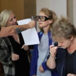 Trzy kobiety biorą udział w aktywności; jedna nosi gogle zniekształcające wzrok, druga trzyma papier, a trzecia dostosowuje okulary, patrząc w dół, co sugeruje demonstrację lub warsztaty.