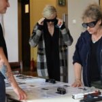 Trzy kobiety stoją wokół stołu z papierami i przedmiotami. Dwie z nich noszą gogle symulacyjne, jedna dostosowuje swoje, podczas gdy trzecia obserwuje. Stół zawiera pomoce dotykowe i materiały informacyjne.
