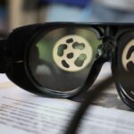 Zbliżenie okularów w czarnych oprawkach spoczywających na papierze, z odbiciem okrągłego wzoru przypominającego czarne plamy na białym tle widocznym w obu soczewkach.