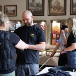 Cztery osoby w czarnych koszulkach Creo wchodzą w interakcję w galerii sztuki. Dwie z nich są zaangażowane w rozmowę, a dwie używają narzędzi do symulacji upośledzenia wzroku, w tym białej laski i gogli. Na ścianach za nimi wyświetlane są dzieła sztuki.