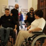 Dwie osoby na wózkach inwalidzkich rozmawiają w pomieszczeniu, podczas gdy inni chodzą i stoją w pobliżu. Otoczenie wydaje się być przestrzenią publiczną lub społeczną z plakatami i oprawionymi zdjęciami na ścianach.