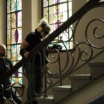 Kobieta w słuchawkach i ciemnych okularach, trzymająca laskę, wchodzi po schodach z ozdobnymi poręczami. Mężczyzna stoi za nią, obserwując, przed kolorowymi witrażami.