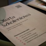 Zbliżenie broszury w języku polskim zatytułowanej Oferta Szkoleniowa z danymi kontaktowymi Stowarzyszenia CREO, umieszczonej na ciemnej powierzchni. Broszura oferuje kursy szkoleniowe dostosowane do indywidualnych potrzeb.