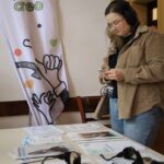Kobieta w słuchawkach stoi przy stole z broszurami i urządzeniami audio. Za nią znajduje się biały baner z kolorowymi postaciami i słowem "creo" oraz ilustracjami dłoni i serc.