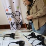 Kobieta w słuchawkach stoi przy stole z kilkoma zestawami słuchawkowymi audio i arkuszami informacyjnymi. Za nią znajduje się baner z kolorowymi postaciami ludzkimi i słowem creo nad zilustrowanymi dłońmi tworzącymi gesty.
