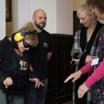 Kobieta ubrana w kombinezon symulacyjny, opaskę na oczy i słuchawki korzysta z laski w asyście trzech obserwujących ją osób, dwóch kobiet i jednego mężczyzny, w pomieszczeniu. Grupa wydaje się być zaangażowana w interaktywną aktywność.