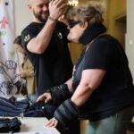 Mężczyzna pomaga kobiecie założyć nakrycie głowy, podczas gdy ona ma na sobie czarny sprzęt ułatwiający poruszanie się i okulary przeciwsłoneczne przy stoliku z napisem REJESTRACJA POCZTOWA. W tle stoi inna osoba.