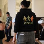 Kilka dorosłych osób wchodzi w interakcję na korytarzu. Jedna z siedzących kobiet nosi zestaw VR i siedzi przy stole z papierami. Inna kobieta stoi w pobliżu, ubrana w bluzę z kapturem creo z kolorowymi postaciami na plecach.