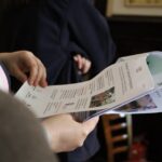 Osoba trzyma i czyta złożony biuletyn lub broszurę w pomieszczeniu, z inną osobą stojącą w rozmytym tle w pobliżu drewnianego krzesła.