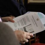 Osoba trzymająca wydrukowane dokumenty, w tym jeden zatytułowany Student Application Form, z widocznym tekstem i małym diagramem. Inna osoba i krzesło z czerwoną poduszką są rozmyte w tle.