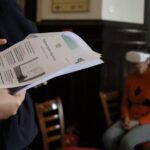 Osoba trzyma wydrukowane dokumenty, podczas gdy inna osoba, siedząca na krześle w tle, nosi biały zestaw słuchawkowy do rzeczywistości wirtualnej i pomarańczowy sweter. Sceneria wydaje się być wewnątrz, prawdopodobnie w klasie lub warsztacie.