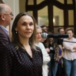 Kobieta stoi przed mikrofonem, przemawiając do publiczności. W tle zgromadzeni są ludzie, niektórzy ze skrzyżowanymi ramionami, a w pobliżu stoi mężczyzna w okularach. Sceneria wydaje się być wewnątrz podczas wydarzenia publicznego.