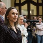 Na pierwszym planie stoi kobieta mówiąca do mikrofonu, a obok niej mężczyzna. W tle zgromadzonych jest kilka osób, które uważnie słuchają w dobrze oświetlonym pomieszczeniu.