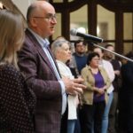 Mężczyzna przemawia do mikrofonu podczas imprezy w pomieszczeniu, obok niego stoi kobieta, a w tle słychać grupę ludzi.