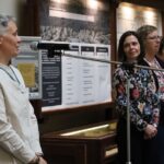 Jedna osoba mówi do mikrofonu, podczas gdy pięć innych stoi w rzędzie obok, słuchając. Znajdują się w pomieszczeniu, przed plakatami informacyjnymi i drewnianą gablotą.
