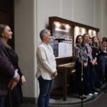 Grupa ludzi stoi w korytarzu wyłożonym gablotami i tablicami, z niektórymi zwróconymi do przodu, a innymi do siebie, co sugeruje formalne lub zorganizowane spotkanie lub wydarzenie.
