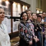 Osoba mówi do mikrofonu, podczas gdy grupa ludzi stoi i słucha uważnie w muzeum lub przestrzeni wystawowej z tablicami wystawowymi w tle.