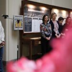 Kobieta mówi do mikrofonu, podczas gdy grupa ludzi stoi i słucha w pobliżu w otoczeniu przypominającym muzeum, z wyświetlaczami informacyjnymi na ścianie i różowymi kwiatami rozmytymi na pierwszym planie.