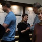 Kilka osób stoi i rozmawia przed tablicami z tekstem i rysunkami architektonicznymi w muzeum lub przestrzeni wystawienniczej. Jedna kobieta uśmiecha się podczas rozmowy, a inna osoba stoi na pierwszym planie i patrzy w kamerę.