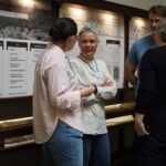 Cztery osoby stoją i rozmawiają przed gablotą i panelami informacyjnymi w muzeum lub na wystawie. Dwie osoby stoją naprzeciwko siebie, podczas gdy inni obserwują i słuchają.