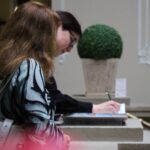 Dwie kobiety stoją przy ladzie. Jedna, na pierwszym planie, z falującymi włosami, stoi bokiem, podczas gdy druga, w tle, pisze długopisem na papierze. Obok nich na ladzie stoi roślina doniczkowa.