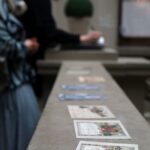 Dwie kobiety stoją w pobliżu długiej kamiennej powierzchni z ilustrowanymi pocztówkami lub małymi wydrukami rozłożonymi na pierwszym planie; jedna kobieta pisze, podczas gdy druga patrzy. Tło i postacie są delikatnie nieostre.