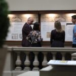 Cztery osoby stoją przed wystawą, czytając informacje na panelach ściennych. Scena rozgrywa się w pomieszczeniu, z balustradą na pierwszym planie i ozdobnymi roślinami w pobliżu.