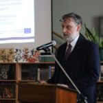 Mężczyzna w garniturze przemawia na podium z mikrofonem w bibliotece. Za nim wyświetlany jest slajd prezentacji na temat platform cyfrowych dla zbiorów specjalnych. W tle widoczne są półki z książkami i schody.