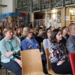Grupa dorosłych siedzi uważnie w rzędach krzeseł w bibliotece, słuchając prezentacji lub wykładu. Regały wypełnione książkami ustawione są wzdłuż ścian w tle.