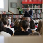 Mężczyzna w garniturze przemawia na podium przed publicznością w bibliotece. Ludzie siedzą, słuchając uważnie, a w tle widoczne są półki z książkami i ekran projektora.