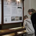 Kobieta i dziecko stoją blisko siebie, czytając wyświetlane informacje i badając dokumenty i artefakty w szklanej gablocie w muzeum lub na wystawie.