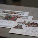 Trzy pocztówki w stylu vintage z odręcznymi wiadomościami i ilustrowanymi punktami orientacyjnymi są ułożone na teksturowanej powierzchni. Pocztówki mają ozdobne obramowania i odręczne pismo w starym stylu.