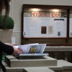 Osoba z długimi włosami stoi przy wystawie, przerzucając strony książki lub broszury w muzeum lub na wystawie z tablicami informacyjnymi w tle.