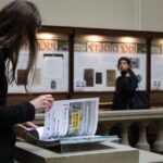 Osoba przegląda dużą broszurę na stojaku w muzeum lub galerii, podczas gdy inna osoba stoi w tle, patrząc na eksponaty ścienne z dokumentami historycznymi i napisem Francia Obli.