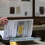 Osoba przeglądająca książkę lub czasopismo, ze stroną przedstawiającą plakat wystawy w języku polskim. W tle widoczne są niewyraźne gabloty z historycznymi dokumentami.