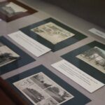 Gablota zawierająca kilka starych czarno-białych i kolorowych pocztówek, każda zamontowana na kolorowym podkładzie z opisowymi etykietami, przedstawiająca historyczne budynki i sceny.