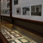 Ekspozycja muzealna ze szklanymi gablotami zawierającymi zabytkowe fotografie i dokumenty oraz panelami informacyjnymi z obrazami i tekstem zamontowanymi na ścianie powyżej.