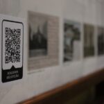 Zbliżenie kodu QR z napisem "Ikonografia Wielkopolska" na ścianie, w tle rozmazane czarno-białe historyczne fotografie.