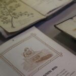 Otwarte książki wystawione na stole, w tym jedna strona z ilustracją osoby siedzącej przy biurku i druga strona z botanicznym rysunkiem gałęzi z liśćmi.