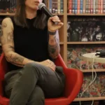 Kobieta z tatuażami na ramionach siedzi na czerwonym krześle i mówi do mikrofonu przed regałem wypełnionym komiksami.